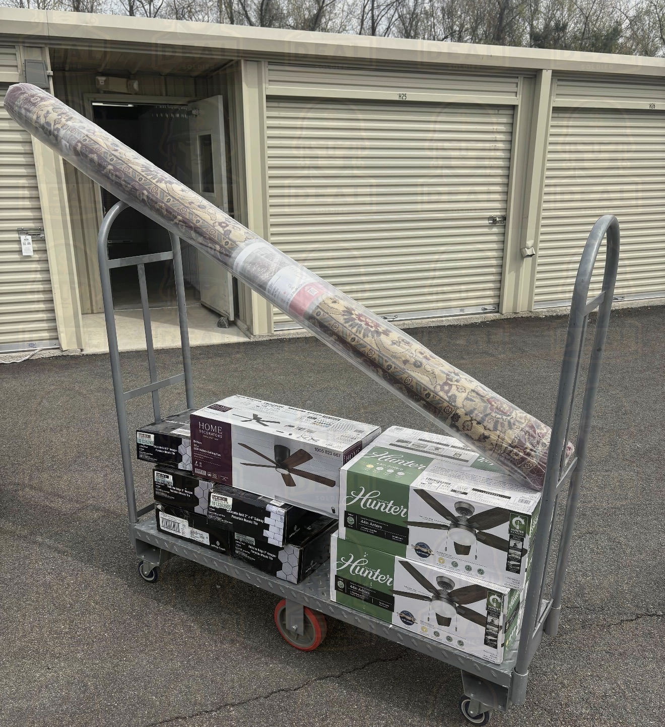 Home Depot flat cart outside a storage unit loaded with a rolled 8x10 rug, three boxed ceiling fans, and stacked tile boxes.